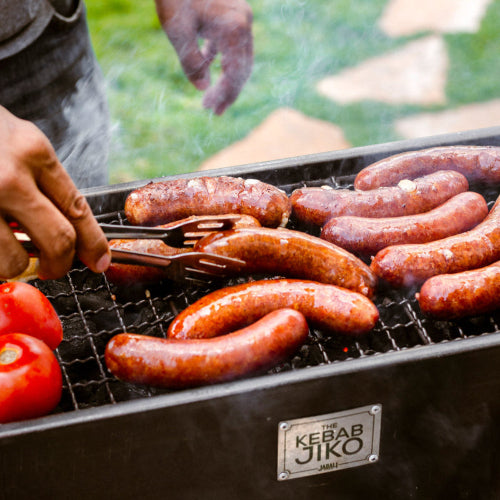 Grilling sausages on a Kebab Jiko grill with a person's hand using tongs.